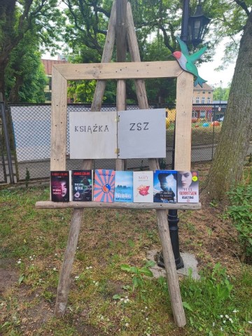 drewniana sztaluga w parku z napisem książka ZSZ na niej siedem książek