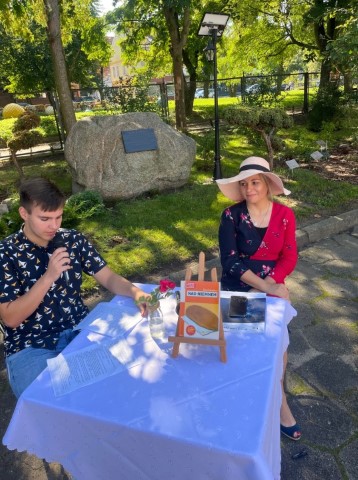 kobieta w kapeluszu i chłopiec z mikrofonem siedzą przy stoliku chłopiec czyta kobieta przygląda się w tle park i kamień